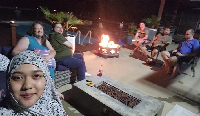 Group of people sitting around a fire pit at night on a patio near a swimming pool, enjoying a relaxed outdoor gathering.
