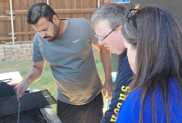 Three people gathered around an outdoor gas grill under a wooden patio cover.