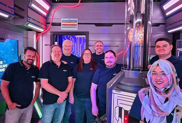 A group of eight people, seven wearing matching dark blue shirts and one woman wearing a light-colored hijab, posing inside a science-fiction themed room.