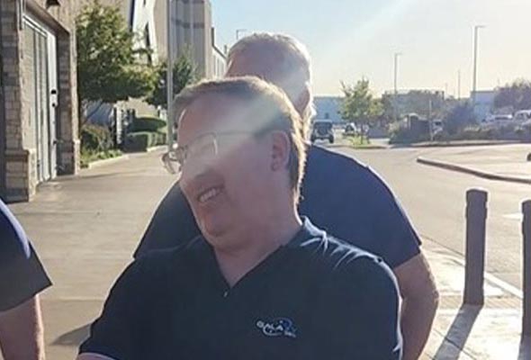 A middle-aged man wearing a dark polo shirt with a light collar and a logo is standing outdoors