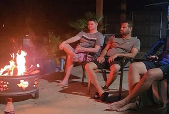 Three men relaxing around a metal fire pit at night