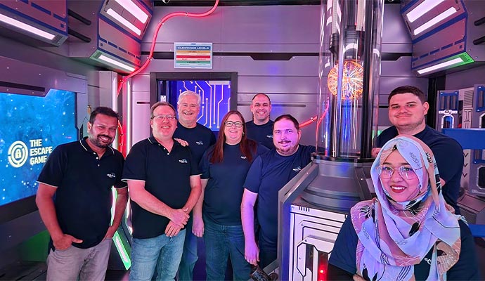 A group of eight team members posing together inside a futuristic escape room with glowing lights and sci-fi themed decor.