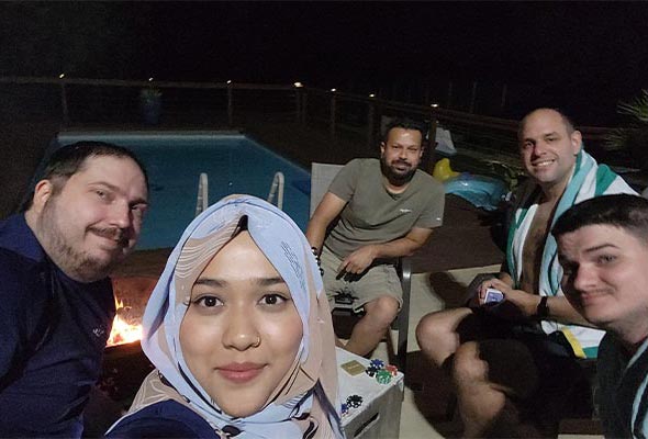A group of friends relaxing by a pool at night, smiling for a selfie near a fire pit.