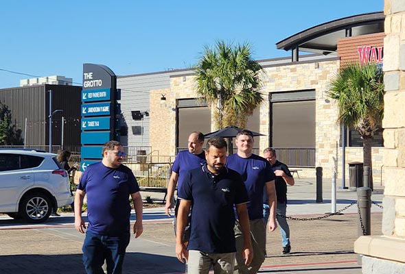 A group of men wearing dark blue polo shirts, are walking toward the viewer on a paved outdoor plaza.