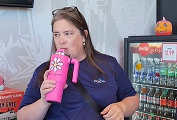 A woman with long hair is standing indoors, drinking from a bright pink tumbler decorated with a white and yellow daisy.