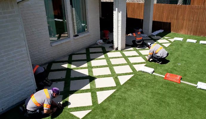 A team of specialized laborers installing synthetic green grass around a diamond-patterned stone patio for a high-end residential backyard