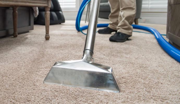 A close-up of a stainless steel carpet cleaning wand on a beige carpet, with the legs of a technician and a blue vacuum hose in the background.