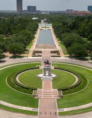 Aerial view of Hermann Park in Houston, Texas, showing the Sam Houston Monument in the foreground, leading to a long reflection pool surrounded by green lawns and trees, with city buildings in the distance