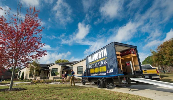 Dalworth Restoration crew at a residential home, with workers carrying a large blue water extraction hose from their truck towards the house. The truck is open, showing drying equipment inside.